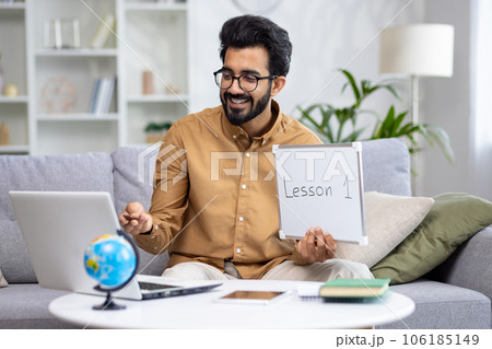 Young male teacher teaching remotely from home, hispanic using laptop for video call and communication and teaching students remotely, explaining new school material smiling. 106185149