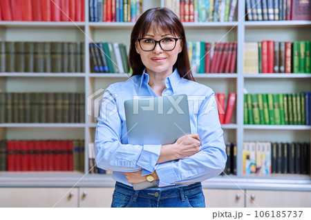 Portrait of confident female teacher holding laptop inside library 106185737