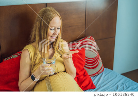 Prenatal Vitamins. Portrait Of Beautiful Smiling Pregnant Woman Holding Pill Box and a glass of water, Taking Supplements For Healthy Pregnancy While Sitting On Couch At Home, Free Space 106186540