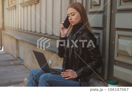 A beautiful caucasian girl with blond hair in a black jacket and blue jeans works with a laptop on the street in clear weather A beautiful caucasian girl with blond hair in a black jacket and blue jeans works with a laptop on the street in clear weather 106187067