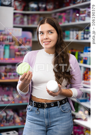 Smiling woman holds two cream for skincare in hands during purchases in cosmetic boutique Smiling woman holds two cream for skincare in hands during purchases in cosmetic boutique 106190379