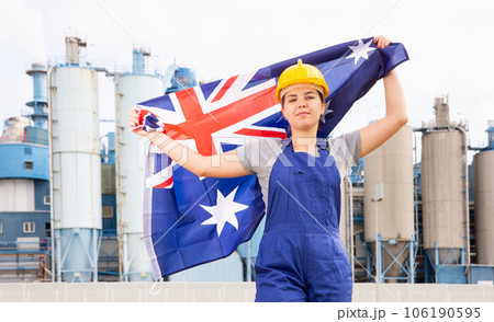 Young female engineer in helmet waving state flag of Australia while standing in front of big tanks at chemical plant Young female engineer in helmet waving state flag of Australia while standing in front of big tanks at chemical plant 106190595
