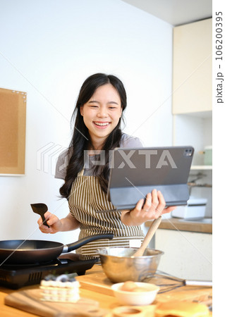 Cheerful asian housewife cooking at home and following an online recipe on digital tablet. People, technology and lifestyle 106200395