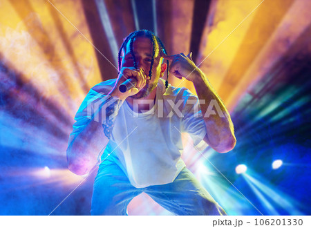 Rap musician. Young artistic man with dreads performing on stage over multicolored neon lights. Concert at night club 106201330
