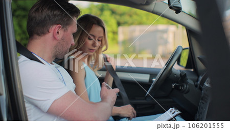 Young woman and driving school instructor with clipboard talking in car. Woman listening to instructor in car learning to drive. 106201555