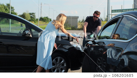 Young caucasian man and woman talking and gesturing looking at broken cars on roadside. Stressed male and female drivers arguing on road accident. 106201685
