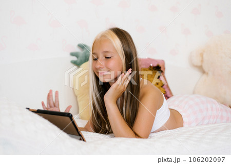 Beautiful teenage girl in pajamas lies on her bed and chatting with her friends and waves hello to them 106202097