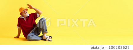 Banner. Flyer. Stylish attractive mature grey-hair man with beard sitting on floor against yellow studio background. Copy space for ad. 106202622