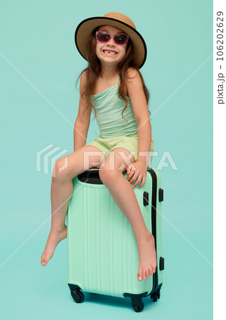 Full length vertical portrait of a Caucasian cute child girl 6 years old, sitting on her suitcase, isolated background 106202629