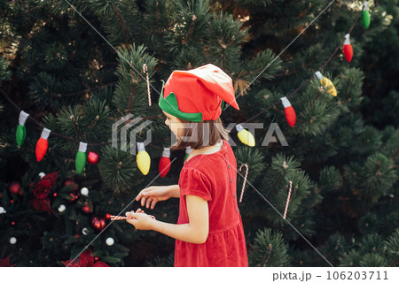 Child waiting for Christmas in wood in summer. portrait of little girl in red dress decorating christmas tree. winter holidays and people concept. Merry Christmas and Happy Holidays Child waiting for Christmas in wood in summer. portrait of little girl in red dress decorating christmas tree. winter holidays and people concept. Merry Christmas and Happy Holidays 106203711