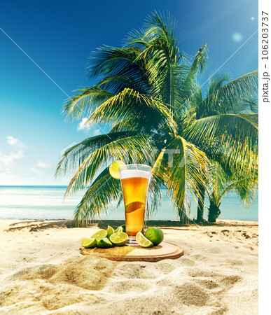 Glass with delicious lager beer with lime appetizers on sand near palm with blue sky and ocean background 106203737