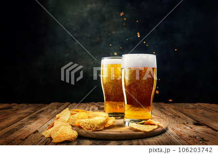 Two glasses of foamy, chill, lager beer with chips appetizers on wooden table against dark background 106203742