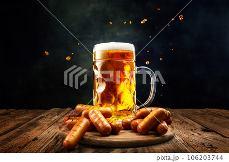Mug with delicious, fresh, foam lager beer with grilled sausages as appetizers on wooden table over dark background 106203744