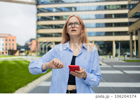 Angry irritated young business woman holding mobile phone and looks at his watch at the blurry business center building 106204374