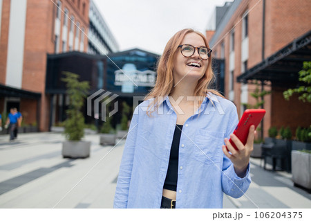 Cheerful happy stylish businesswoman using smartphone online app while standing near the mall. Happy woman in a blue shirt walks down the street looking at her smartphone.An exciting application used 106204375