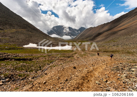 Several backpackers walk along a rocky mountain trail. Big mountains in the background in the clouds. Not melted snow on the ground. Horizontal. Several backpackers walk along a rocky mountain trail. Big mountains in the background in the clouds. Not melted snow on the ground. Horizontal. 106206166