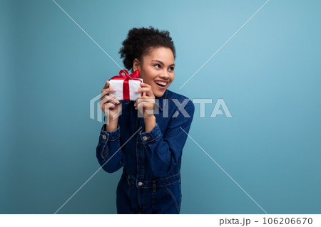a slender young hispanic brunette woman with fluffy curly hair in a blue denim suit hugging a gift a slender young hispanic brunette woman with fluffy curly hair in a blue denim suit hugging a gift 106206670