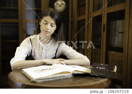 Beautiful girl is sitting at table with a book against the background of a bookcase and a wall clock Beautiful girl is sitting at table with a book against the background of a bookcase and a wall clock 106207219