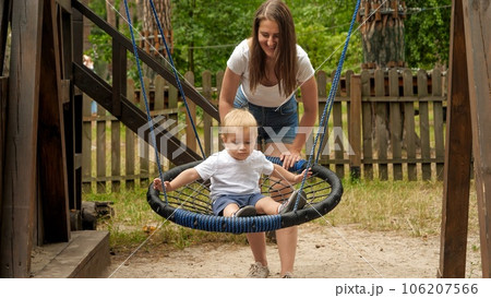 Young mother pushing her baby son swinging in rope nest swing in park. Kids playing outdoors, children having fun, summer vacation Young mother pushing her baby son swinging in rope nest swing in park. Kids playing outdoors, children having fun, summer vacation 106207566