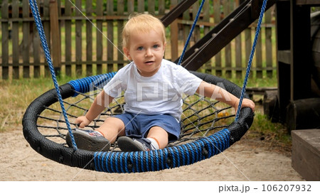 Cute smiling toddler boy swinging in rope nest swing at park 106207932