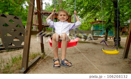 Little boy smiling as swinging on swing at adventure park. Kids playing outdoors, children having fun, summer vacation and holiday. 106208132