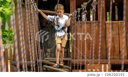 Little boy tied to safety rope crossing wobbly rope bridge at summer adventure park. Kids sports, summer holiday, fun outdoors, scouts. 106208191