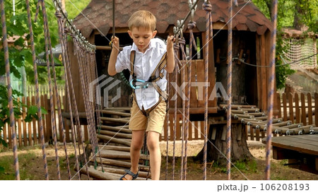 Portrait of brave boy crossing bridge made of planks and ropes at extreme adventure park. Kids sports, summer holiday, fun outdoors, scouts. 106208193