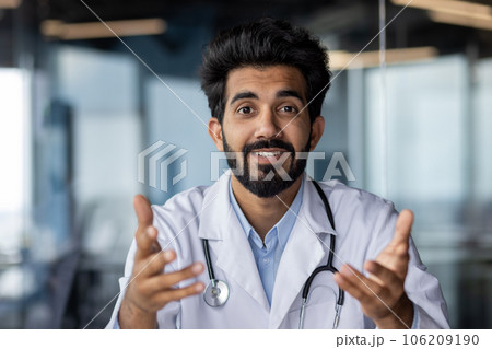 Portrait of a young Indian male doctor, psychologist who conducts online consultation with patients. Smiling, looking at the camera, gesturing with his hands, sitting in the office in the clinic. Portrait of a young Indian male doctor, psychologist who conducts online consultation with patients. Smiling, looking at the camera, gesturing with his hands, sitting in the office in the clinic. 106209190