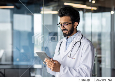 A young smiling Indian doctor works at a tablet, stands in the lobby of the clinic in a white coat. 106209198