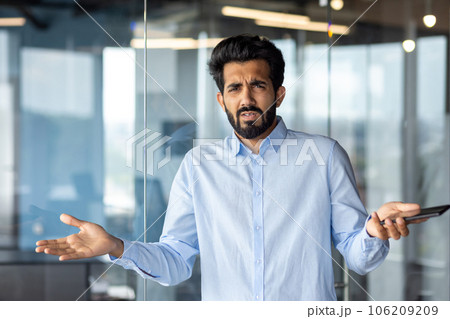 Upset young Indian male businessman standing in office with phone in hand, spreading hands and looking disappointed at camera. 106209209