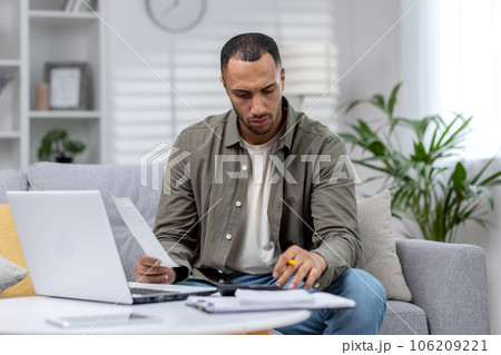 Latin American man is focused working remotely at home on a laptop and with documents. 106209221