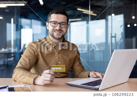 Portrait of a young man working in the office using a laptop, holding a credit card and looking at the camera with a smile. 106209223