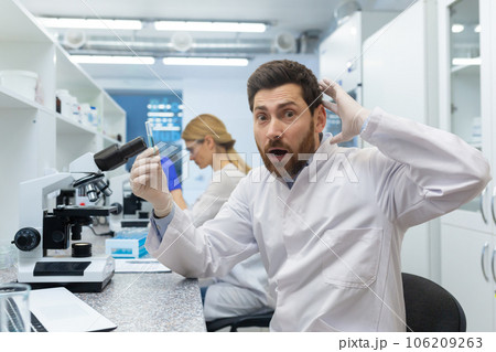 Portrait of a shocked young male scientist sitting in a laboratory behind a microscope, doing research and making a discovery. He holds the substance in his hand and is surprised by the result. Portrait of a shocked young male scientist sitting in a laboratory behind a microscope, doing research and making a discovery. He holds the substance in his hand and is surprised by the result. 106209263