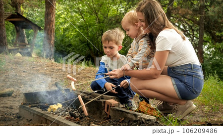 Young mother with two sons cooking sausages on bonfire in camp. Active leisure, children in camping, family vacation in nature. Young mother with two sons cooking sausages on bonfire in camp. Active leisure, children in camping, family vacation in nature. 106209486