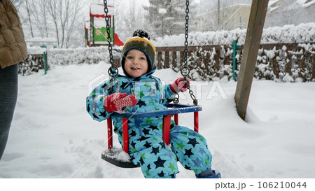 Happy smiling toddler boy enjoying swinging in swing during snowfall at winter park. Fun and joy on winter holidays, kids playing outdoors, activity in snowfall. 106210044