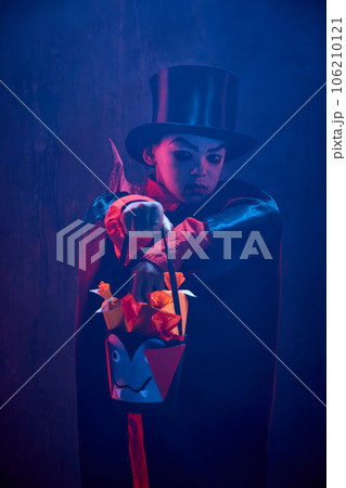 Little boy, child in image of creepy vampire costume and makeup standing with basket for sweets against dark background in neon light with smoke 106210121