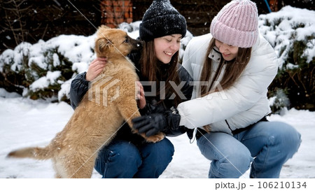 Two happy girls with dog having fun in snow at house backyard. Kids with animals, games with pets. Two happy girls with dog having fun in snow at house backyard. Kids with animals, games with pets. 106210134