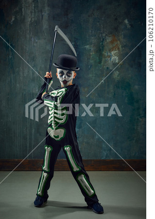Fantasy. Little boy, child with pale face makeup, wearing costume of skeleton, standing with scythe against vintage green background 106210170