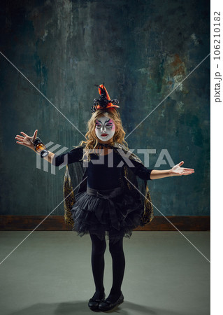 Little girl, child with colorful witch makeup and costume standing against dark vintage background. Mystery 106210182