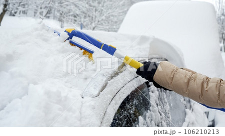Close-up of a woman using a snow brush to remove snow from her car's windshield after a heavy snowfall. Concept of winter weather, car maintenance, and being prepared for snowy conditions. 106210213