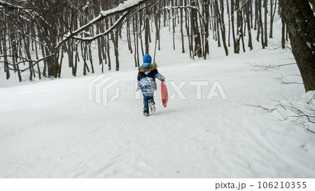 Happy boy is carrying his plastic sleds up the hill, excited for the snow and all the outdoor activities it brings during the winter season. Happy boy is carrying his plastic sleds up the hill, excited for the snow and all the outdoor activities it brings during the winter season. 106210355