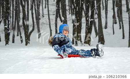 Cheerful young boy rides down a snowy slope on his red plastic sled, his face lit up with excitement and laughter. Winter playtime and the unbridled enthusiasm of youth. Cheerful young boy rides down a snowy slope on his red plastic sled, his face lit up with excitement and laughter. Winter playtime and the unbridled enthusiasm of youth. 106210366
