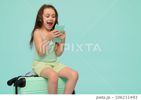 Cheerful little traveler child girl sitting on her blue suitcase, smiles while checks new mobile app on her smartphone Cheerful little traveler child girl sitting on her blue suitcase, smiles while checks new mobile app on her smartphone 106211493