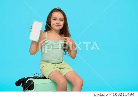 Adorable little traveler girl on a plastic blue valise, showing at camera a smartphone with copy space for mobile app Adorable little traveler girl on a plastic blue valise, showing at camera a smartphone with copy space for mobile app 106211494