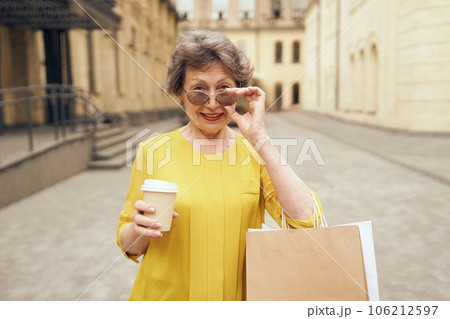 Happy mature woman in sunglasses walking with her shopping bags and coffee cup in city street 106212597