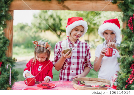 Happy children preparing for Christmas. Two boys and girl in santa hat color gingerbread cookies, drinking hot chocolate outside having fun. kids decorate gingerbread man. Holidays concept Happy children preparing for Christmas. Two boys and girl in santa hat color gingerbread cookies, drinking hot chocolate outside having fun. kids decorate gingerbread man. Holidays concept 106214014