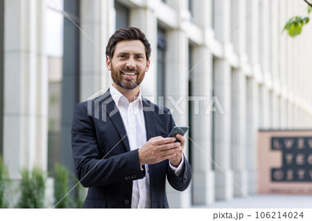 Young business man, office worker standing outside on the street in a suit and using a mobile phone. Smiling at the camera. Young business man, office worker standing outside on the street in a suit and using a mobile phone. Smiling at the camera. 106214024