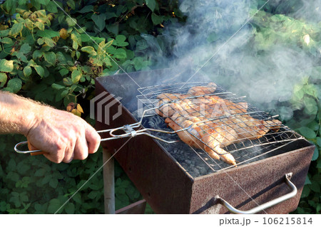 man roasts chicken on an outdoor grill in garden. A close-up of hand turning over grate with meat 106215814