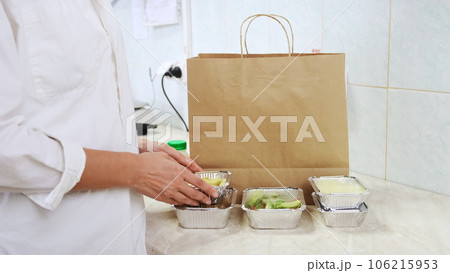 Food delivery service in restaurant. Chef packs lunch boxes to paper bag. Healthy diet daily menu, food meal dishes in a plastic takeaway containers. Woman cook on white kitchen collecting food box Food delivery service in restaurant. Chef packs lunch boxes to paper bag. Healthy diet daily menu, food meal dishes in a plastic takeaway containers. Woman cook on white kitchen collecting food box 106215953