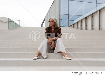 a well-groomed middle-aged woman in glasses dressed in a brown jacket sits on the porch of a a well-groomed middle-aged woman in glasses dressed in a brown jacket sits on the porch of a 106219064